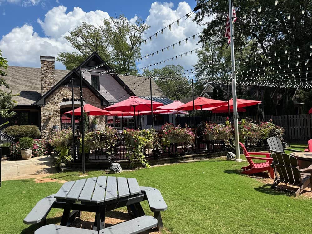Outdoor seating area of a restaurant with red umbrellas, string lights, and a well-maintained lawn with picnic tables and Adirondack chairs in the foreground. Stone building and shrubbery in the background.