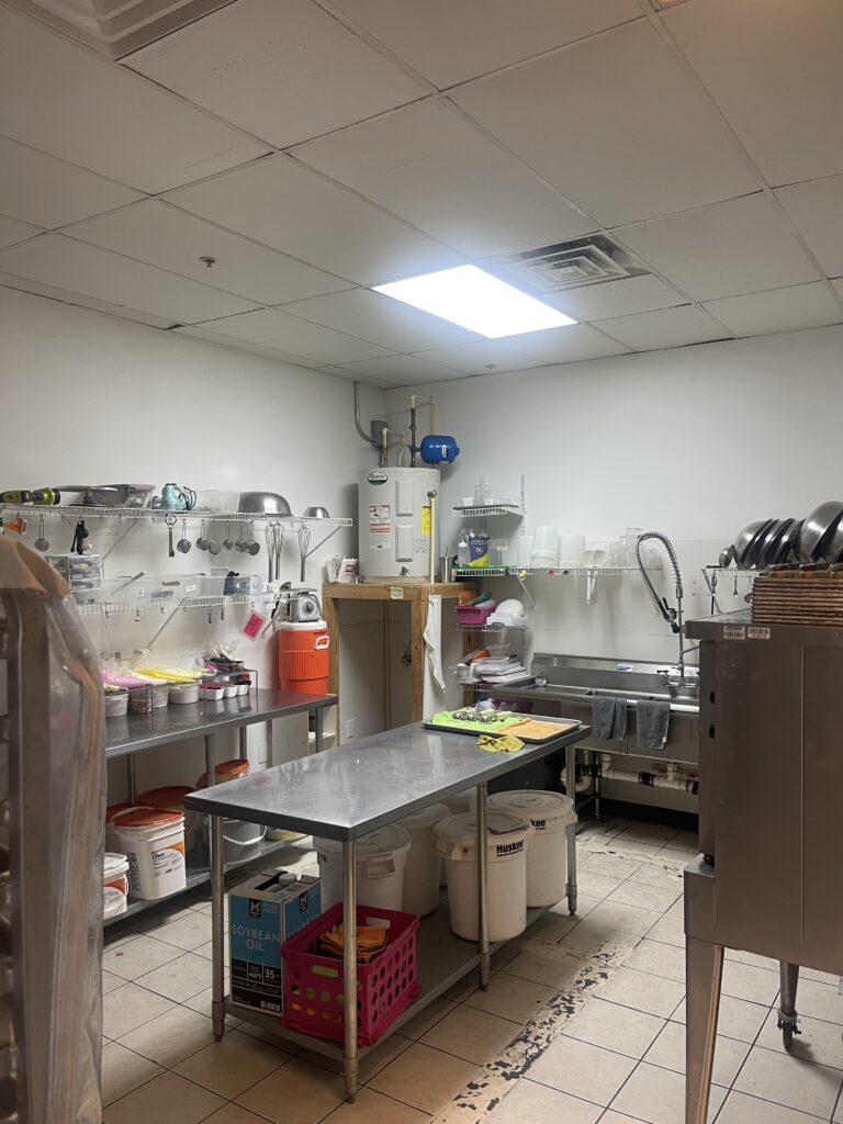 A commercial kitchen in Alpharetta, GA, features stainless steel counters and shelves laden with cookware. Plastic containers and a water heater complete the setup. A sink is on the right, with cleaning supplies neatly stored on the floor, perfect for a bakery for sale seeking street visibility.