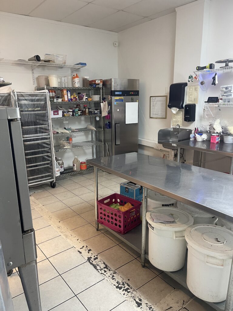 This Avalon commercial kitchen boasts metal tables, shelves stocked with supplies, a refrigerator, and a cart with trays. Floor tiles gleam underfoot, while cleaning items line the right wall. Ideal for an Alpharetta GA Bakery, it offers great street visibility for attracting customers.