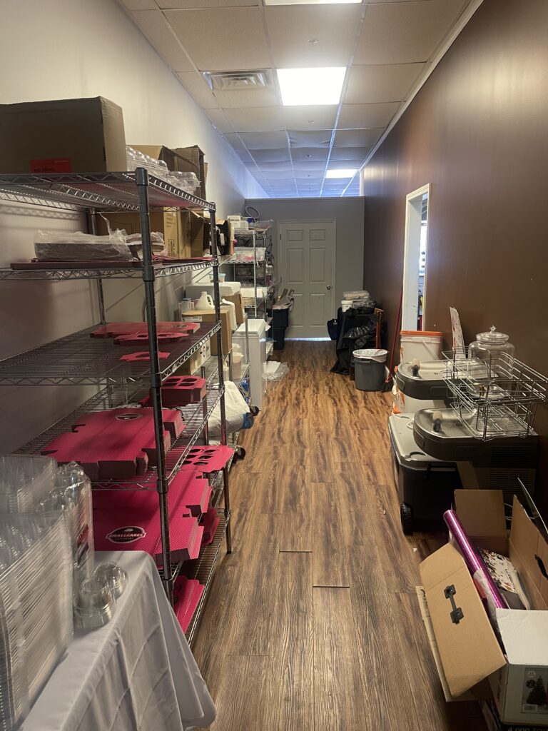 Narrow storage room with metal shelves holding boxes and supplies, reminiscent of a quaint Alpharetta GA bakery. Wooden floor, door at the far end offers great street visibility.