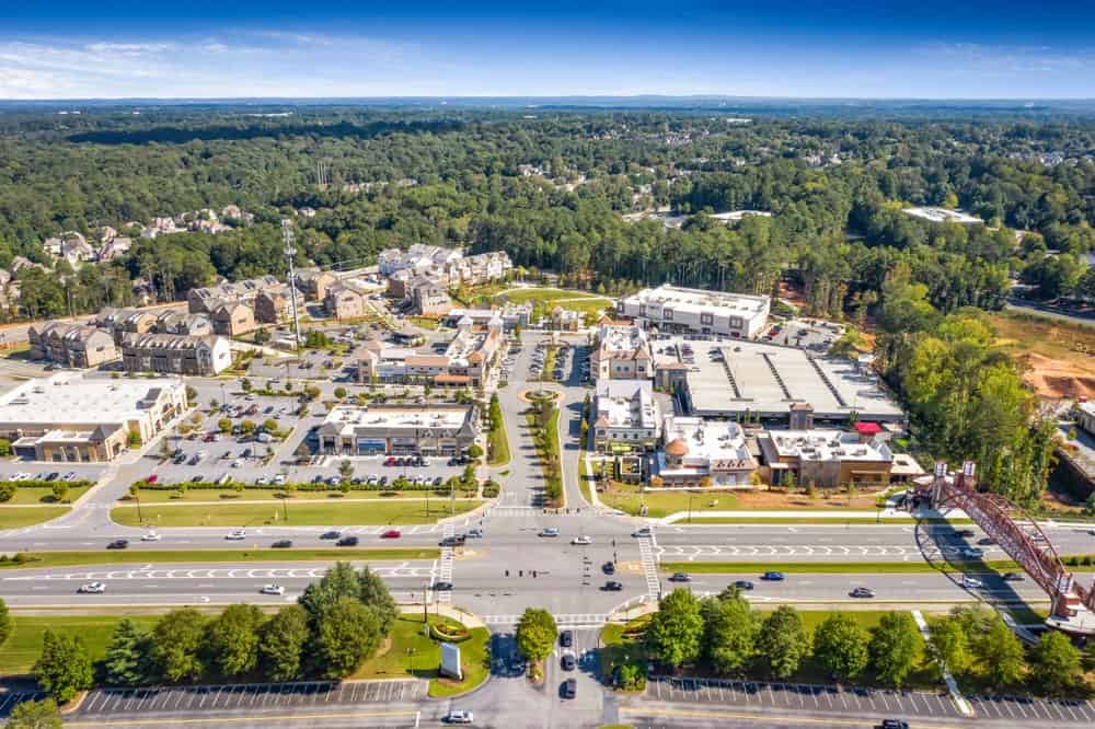 Aerial view of a suburban shopping center with parking lots, roads with cars, and surrounded by trees and residential areas.