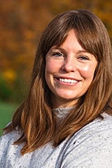 Woman smiling outdoors with long hair, wearing a gray sweater, in front of a blurred natural background.