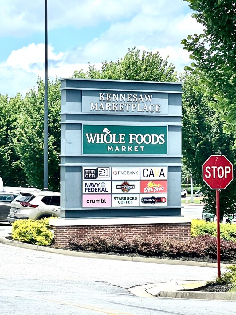 A sign at Kennesaw Marketplace lists Whole Foods Market and several other businesses, including Starbucks, Navy Federal, and Crumbl, with parked cars and greenery in the background.