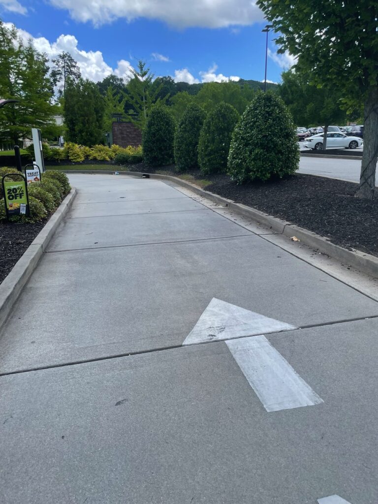 A concrete drive-thru lane at a freestanding restaurant in Kennesaw GA, with a white arrow on the ground, lined by green bushes and trees, under a partly cloudy sky.