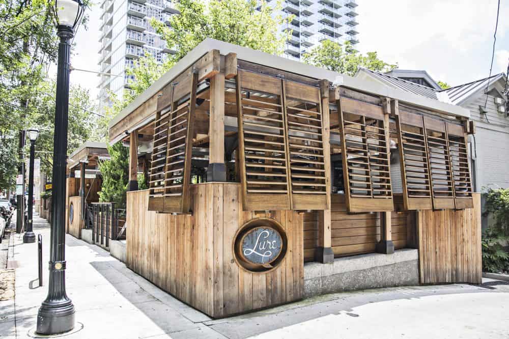 A modern wooden restaurant with large slatted panels on a city street corner, featuring a round sign with "Line" written on it and high-rise buildings in the background.