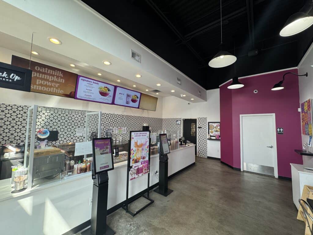 A brightly lit smoothie shop interior with self-service kiosks, a service counter, digital menu screens, and patterned wall tiles.