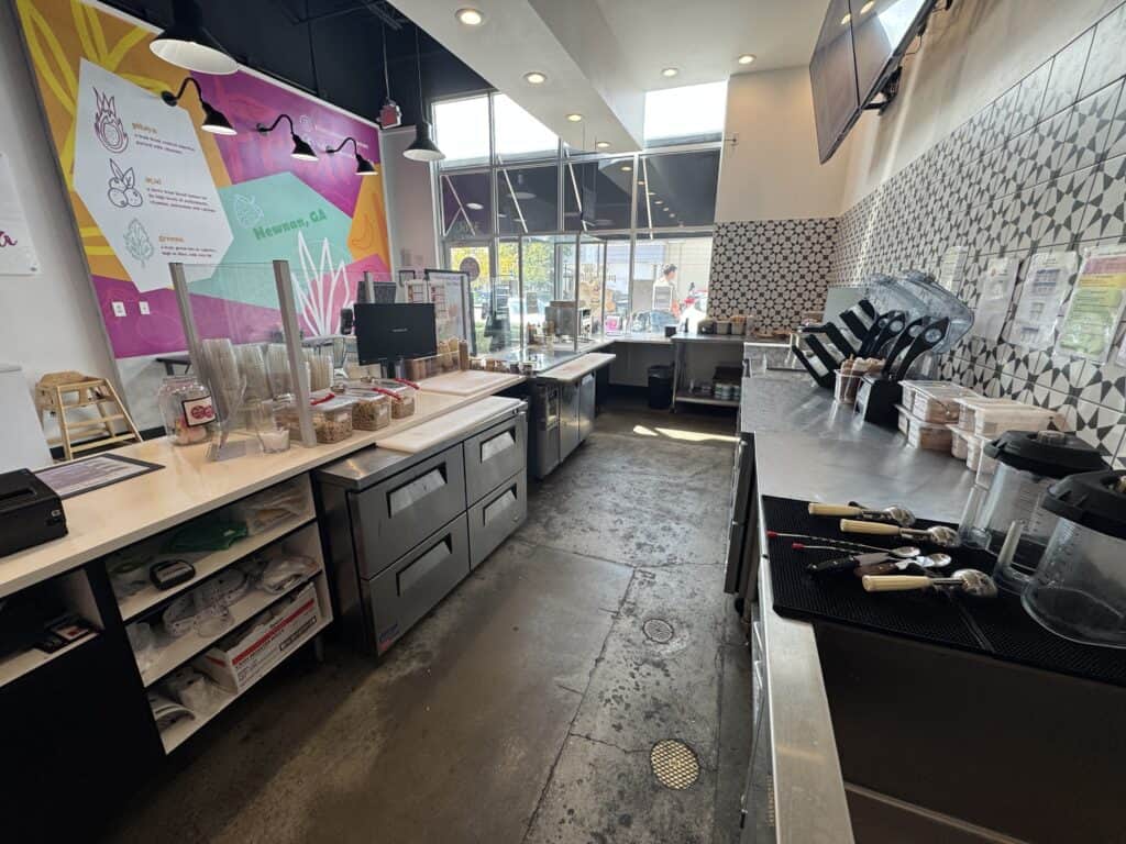 Interior of a modern bubble tea shop with counters, drink machines, utensils, and a colorful wall menu near large front windows.