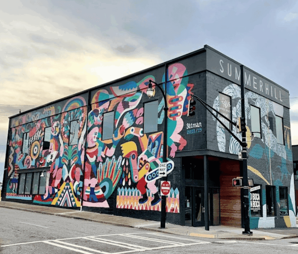 A colorful mural covers the exterior of a corner building labeled “SUMMERHILL,” featuring abstract and animal-like designs near a street intersection with traffic signs, home to a unique Summerhill Atlanta restaurant.