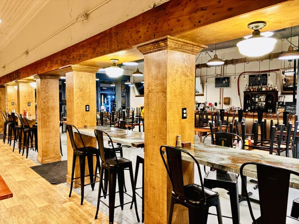 Restaurant interior with wooden pillars, high tables, and stacked chairs, suggesting the place is closed or being cleaned. Bright lighting and bar area visible in the background.