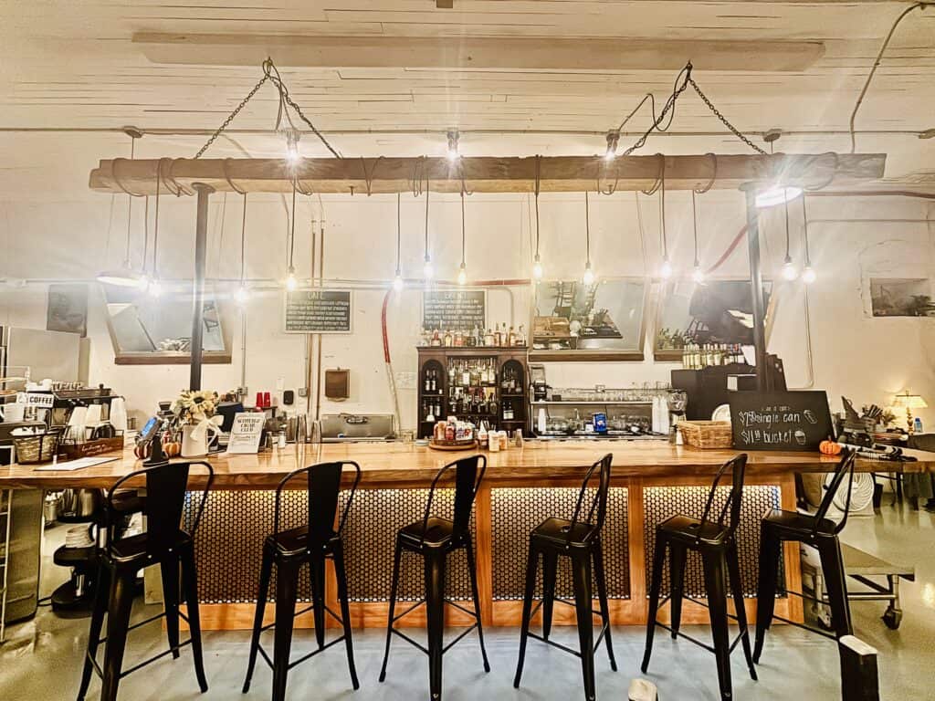 A modern café bar with metal barstools, hanging lights, a wooden counter, and shelves stocked with bottles and coffee-making equipment. Handwritten chalkboard menus are displayed behind the bar.