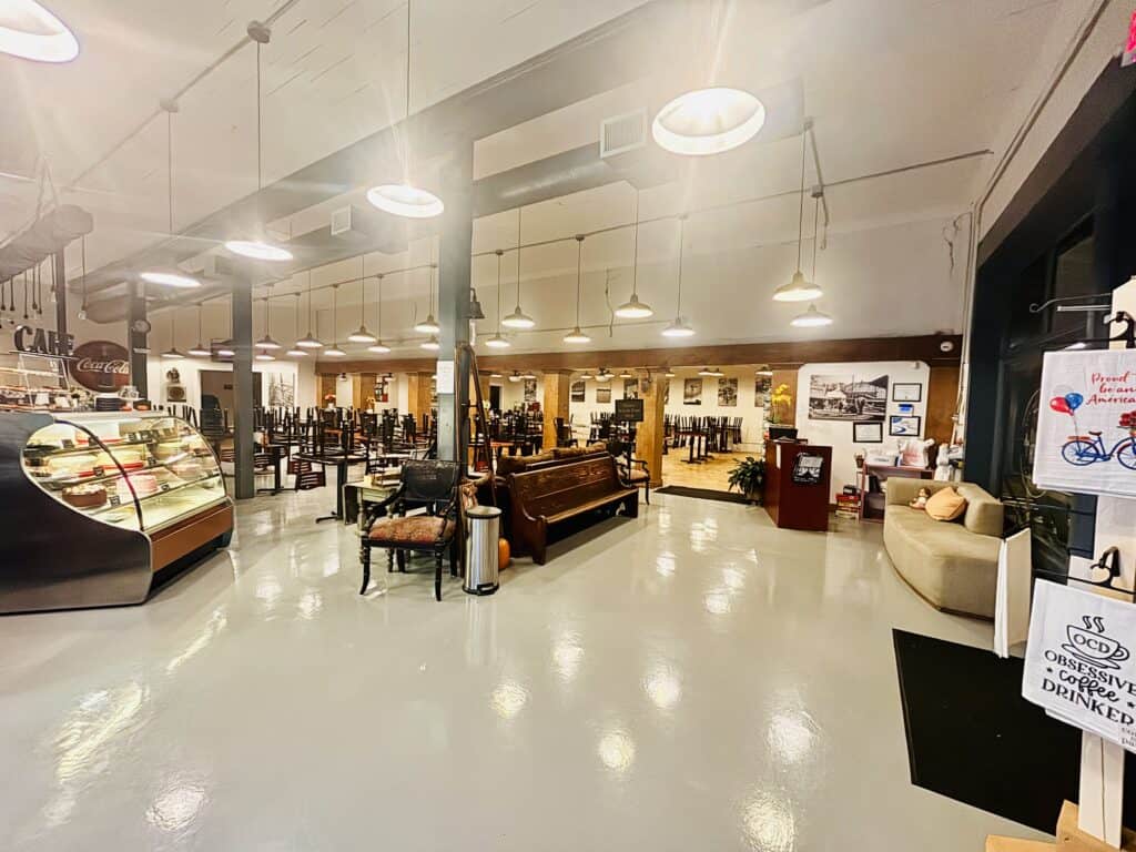 Spacious, well-lit café interior with a pastry display case, seating area, vintage decor, and signs on the walls. Tables and chairs are arranged neatly in the background.