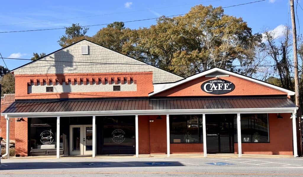 A red brick building with a black awning houses the Concord Cafe, with a sign above the entrance and trees in the background.