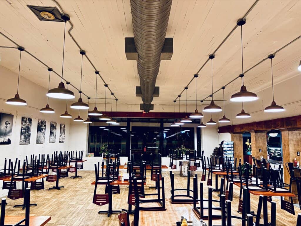 Empty restaurant interior at night with chairs upside down on tables, pendant lights hanging from the ceiling, and large windows at the back.