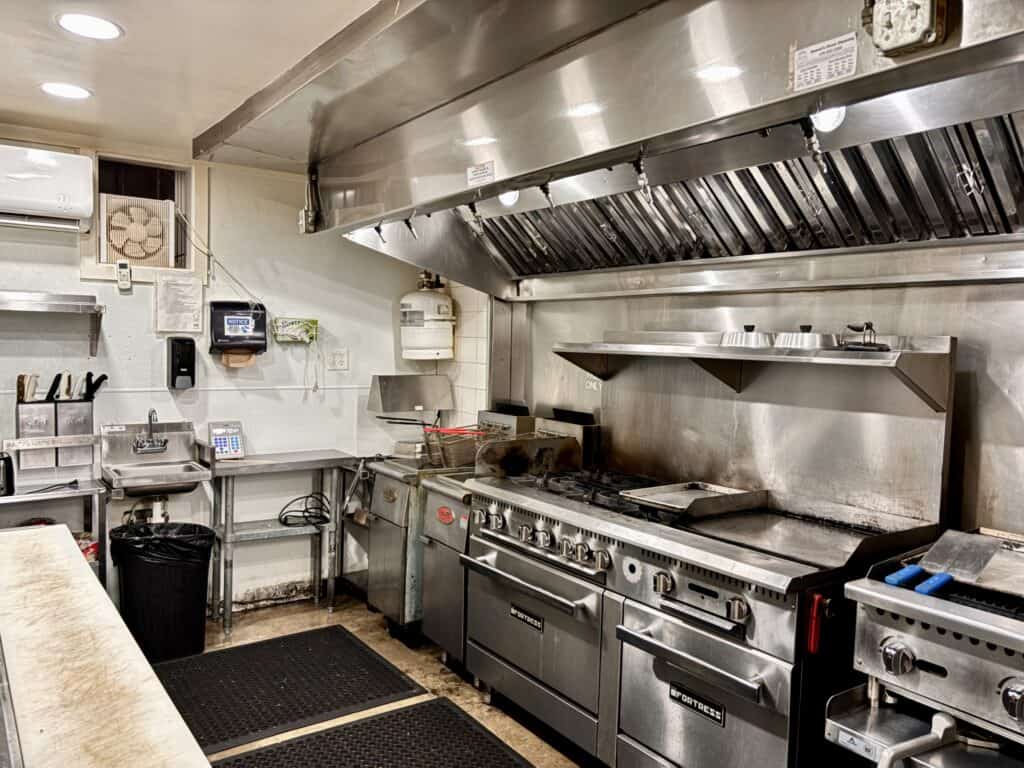 Commercial kitchen with stainless steel appliances, range, fryers, griddle, shelves, and prep area; clean and organized workspace.