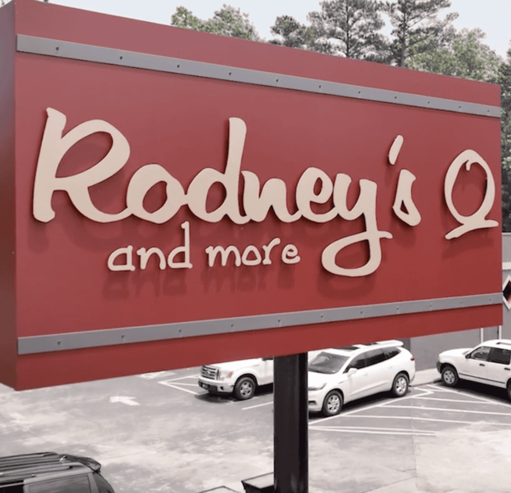 A red sign reads "Rodney's Q and more" above a parking lot with several cars and trees in the background.
