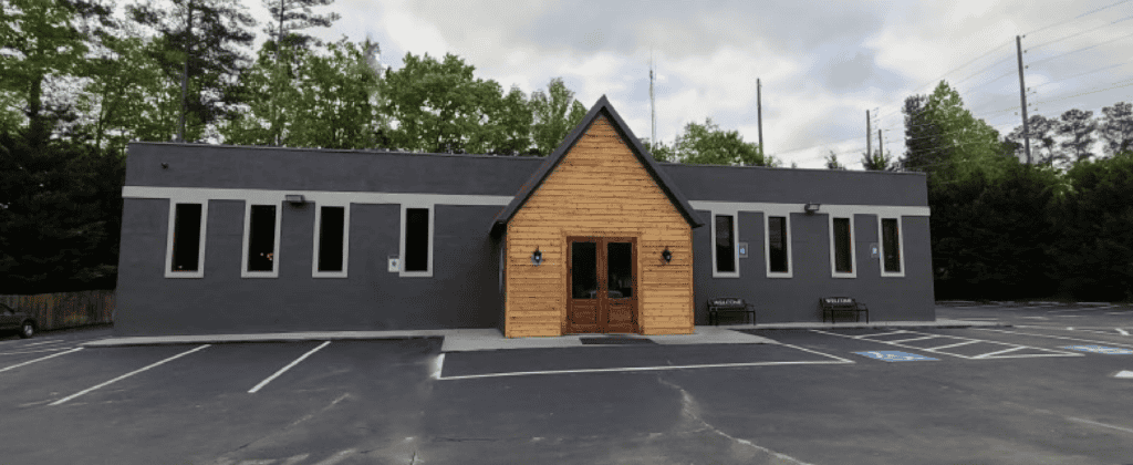 A single-story commercial building with a gray exterior and a wooden entrance, surrounded by trees and a parking lot with marked accessible spaces.