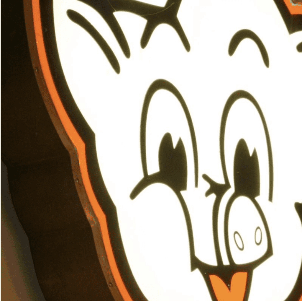 Close-up of a brightly lit cartoon pig face sign featuring large eyes, a round snout, and an orange tongue.